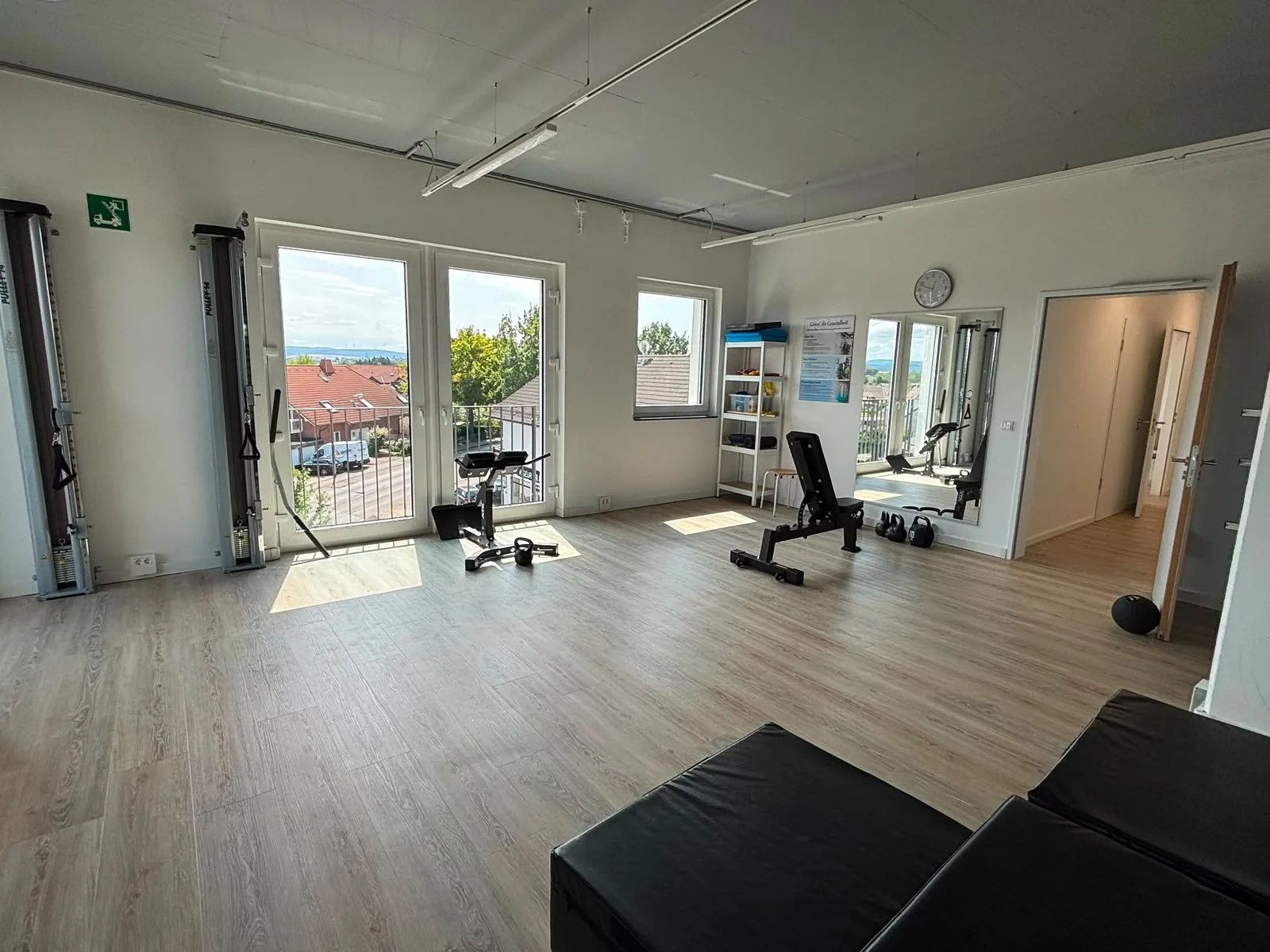 Weite Trainingsfläche mit Seilzügen, Hantelbank und Spiegelwand, Fensterfront über den Dächern des Ortes